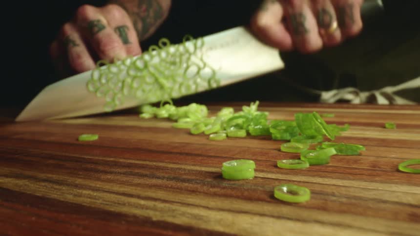 Close up of a tattoed man chopping chives with a sharp kitchen knife over a wooden board