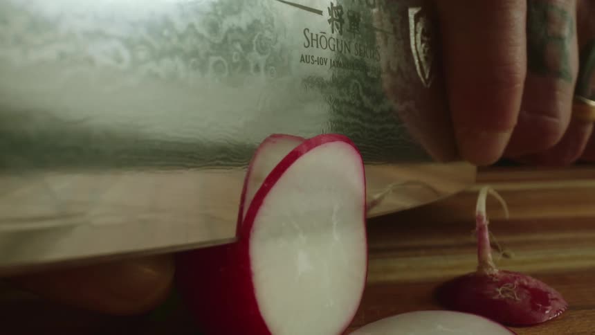 Close up of a tattoed man cutting radishes with a sharp kitchen knife over a wooden board