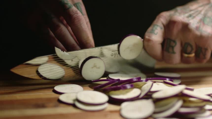 A man with tattoos on his fingers slices a zucchini with a large knife on a cutting board