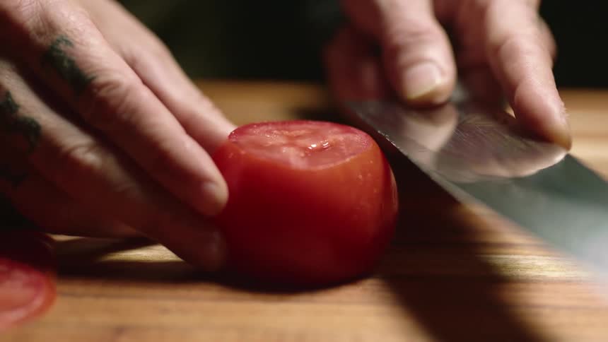 A tattoed man slicing a tomato with a sharp kitchen knife over a wooden board