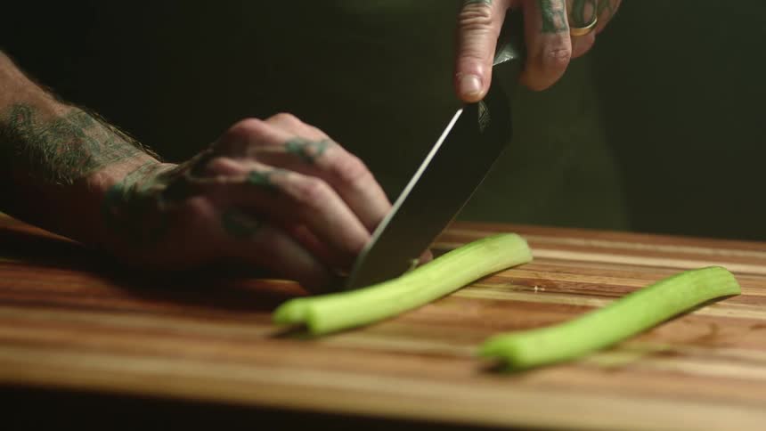 A man with tattoos on his hands and fingers slices a stalk of celery on a wooden cutting board