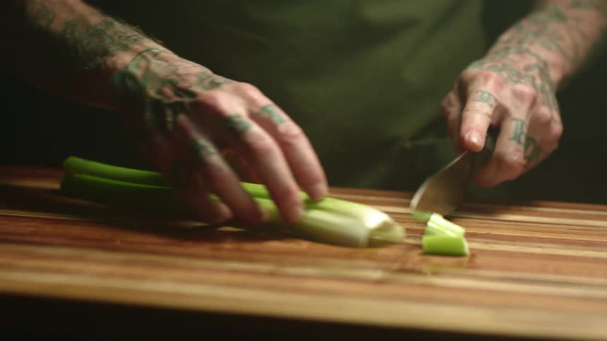 A man with tattooed hands and arms cutting a piece of celery on a wood cutting board