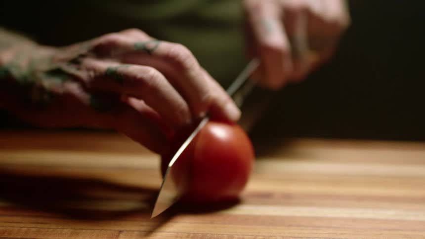Close up of a tattoed man cutting tomatoes with a sharp kitchen knife over a wooden board