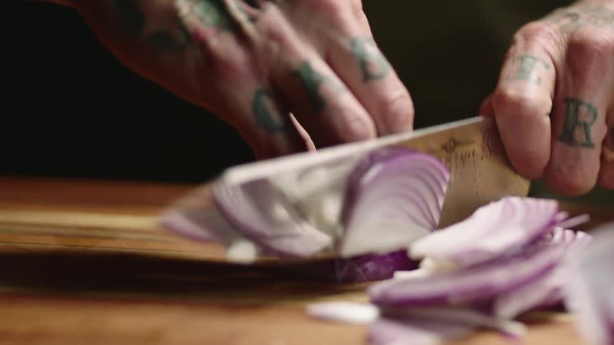 Close up of a tattoed man slicing purple onions with a sharp kitchen knife over a wooden board