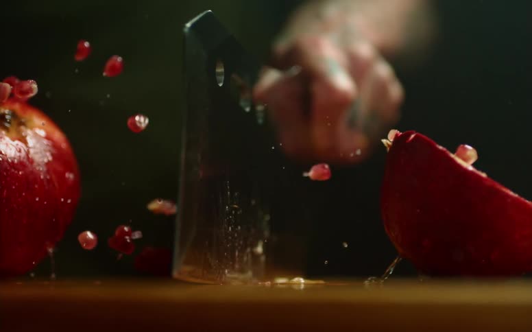 Close up of a tattoed man cutting a pomegranate with a sharp cleaver over a wooden board