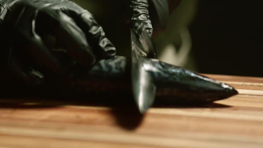 A person wearing black latex gloves gutting a fish with a sharp kitchen knife over a wooden cutting board