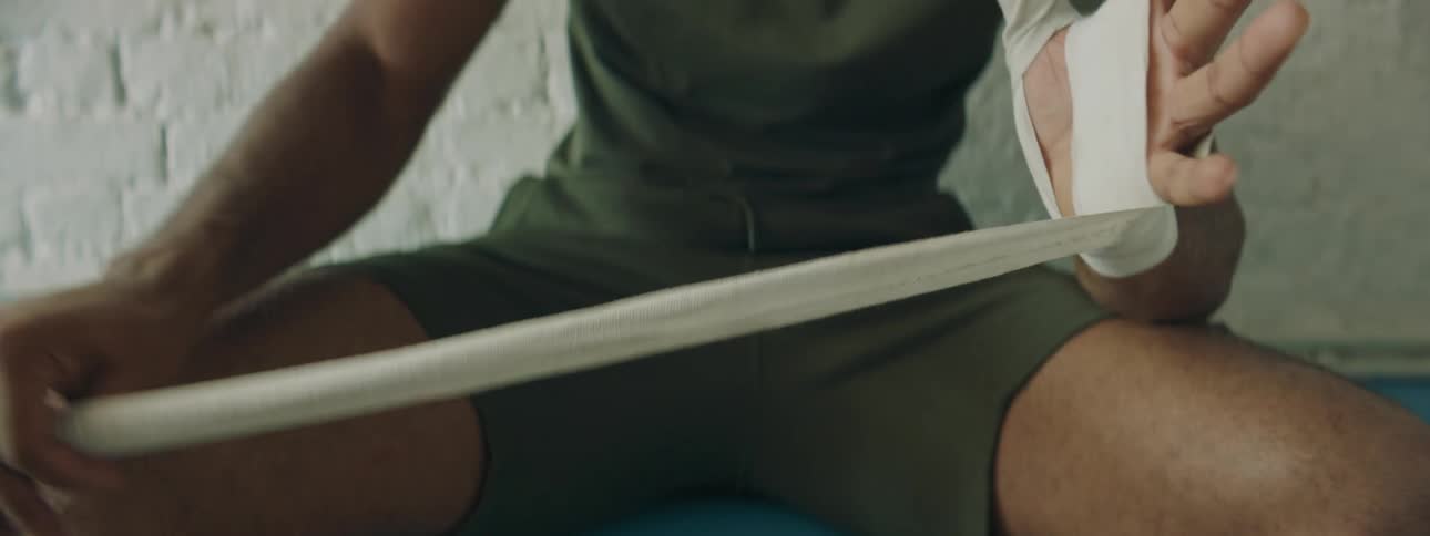 A man wraps his hand with white bandage while sitting in workout clothes with arm muscles bulging
