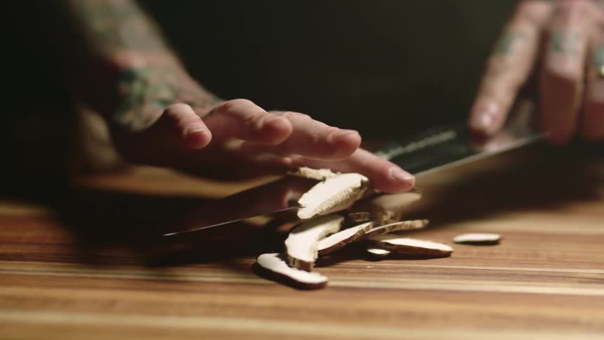 A person with tattooed arms cutting mushrooms on a wood cutting board