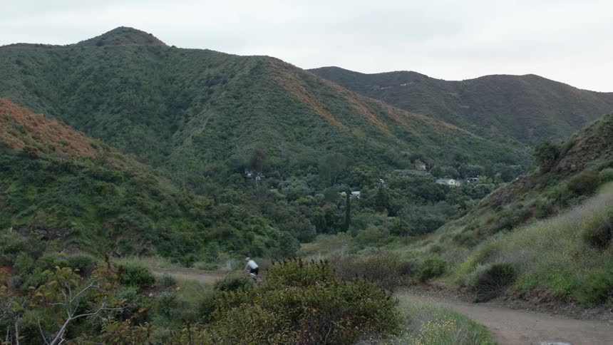 A cyclist rides a bicycle along a dirt road at the base of a verdant mountain range during the day