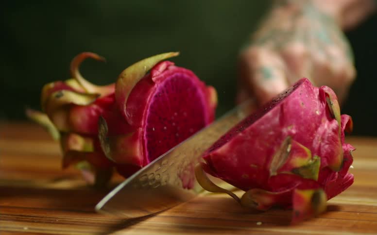 Close up of a tattoed man cutting a dragon fruit with a sharp kitchen knife over a wooden board