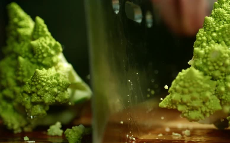 Close up of a tattoed man chopping a romanesco cauliflower with a sharp cleaver over a wooden board