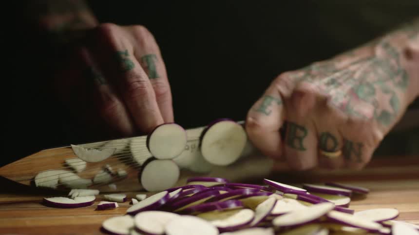 A person with tattooed hands uses a knife to chop vegetables on a wooden cutting board