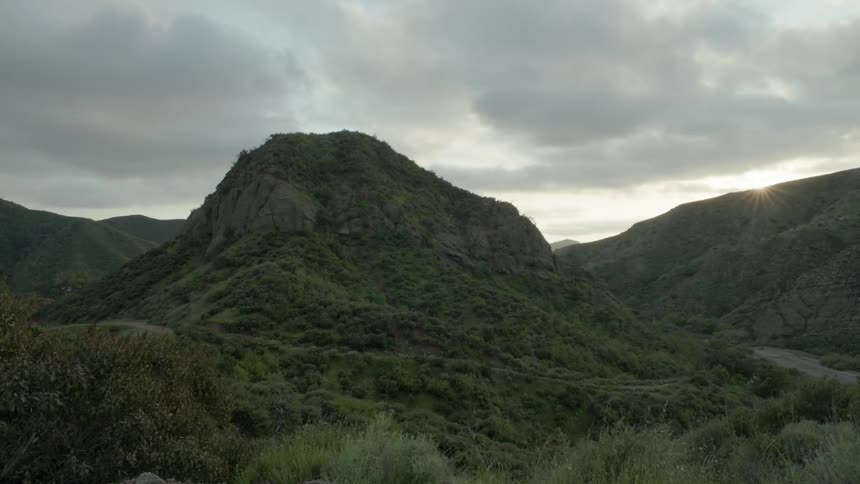 The sun slowly sets over a hilly and mountainous terrain on a cloudy day