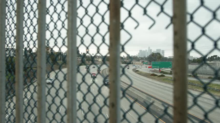 A hole in a chainlink fence on an overpass overlooking a multi lane highway with a city skyline in the distance