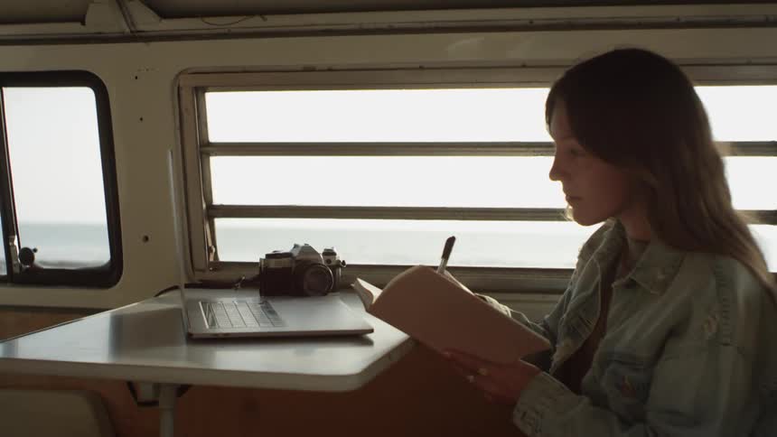 A woman scrolling on a laptop and writing in a notebook while sitting at a table