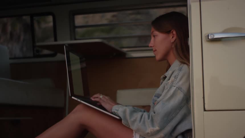A woman types on the laptop resting on her knees as she sits on the floor of a large van during the day