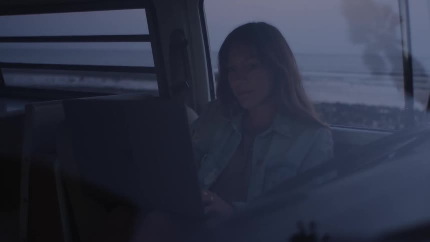 A teenage girl sits in her van parked on the beach and types on her laptop