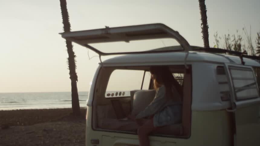 A young woman types on a laptop while sitting in the back of a van parked on the coast during the day