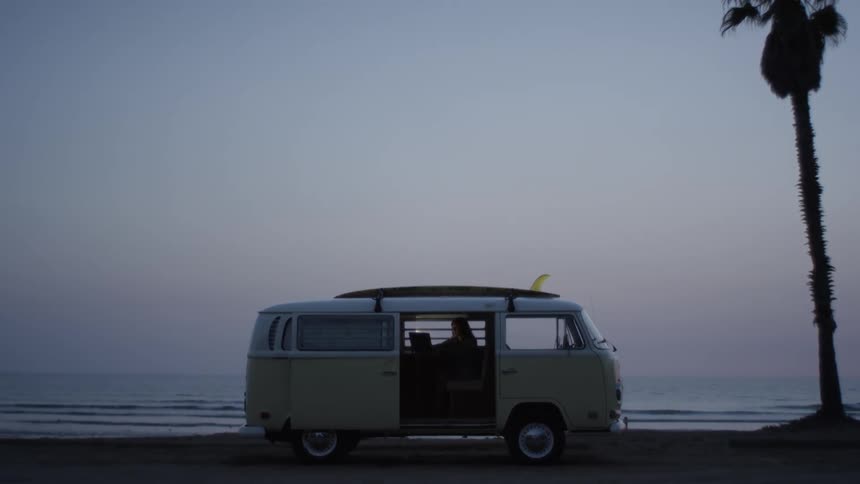 A van parked on a sandy beach next to a silhouetted palm tree at dusk