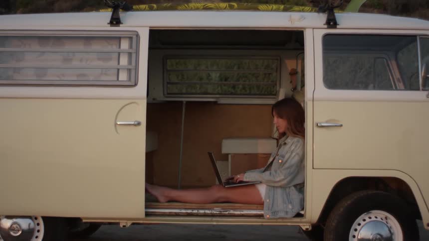 Young woman sitting on the floor of a volkswagen van parked on a rural road working on a laptop