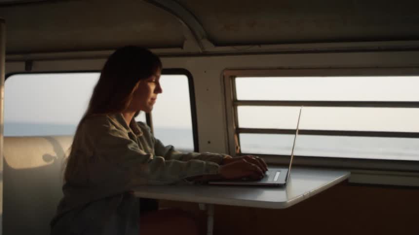 A woman is sitting down and typing on a laptop in an RV during the day near the ocean