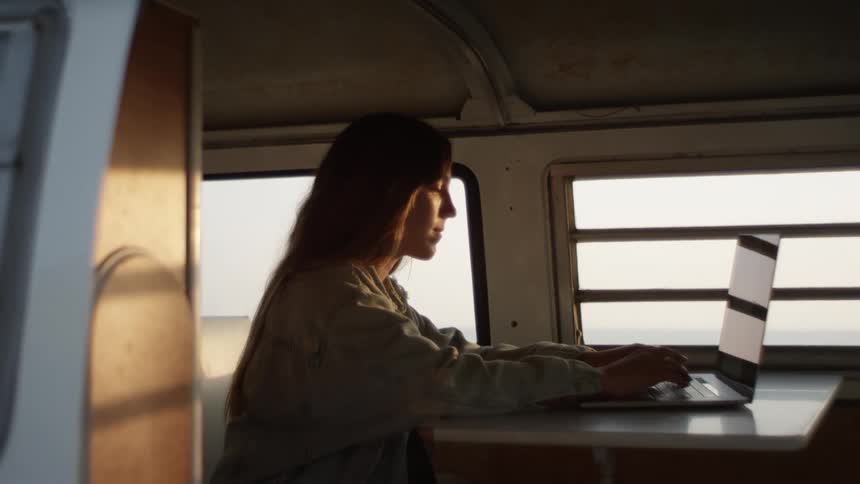 A woman is typing on a laptop next to a window at sunset