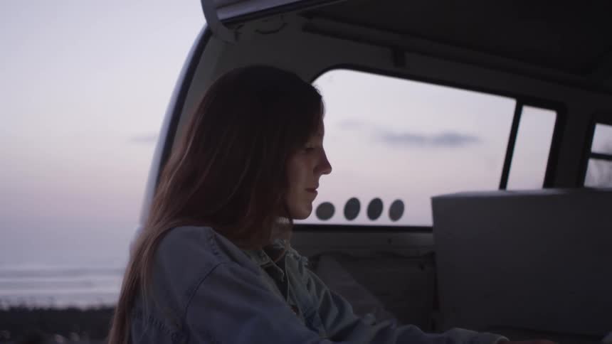 A woman stands at the back of her van at the beach at dusk