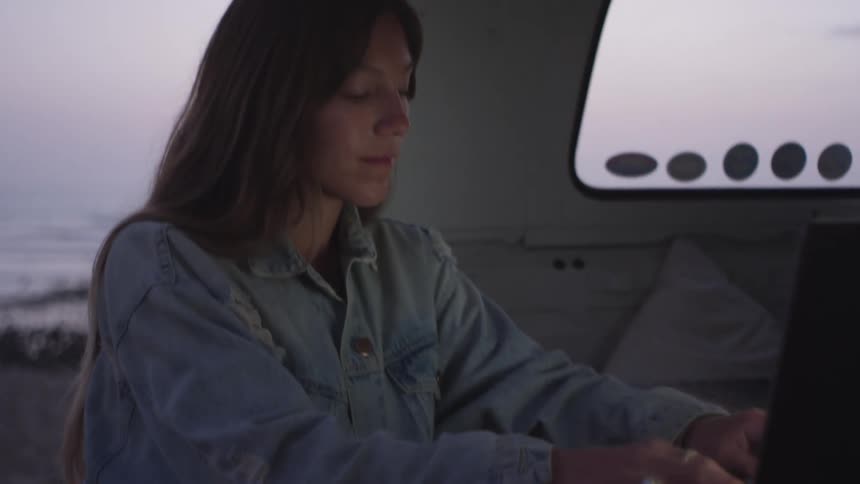 A woman types on her laptop while standing at the back of her van on the beach