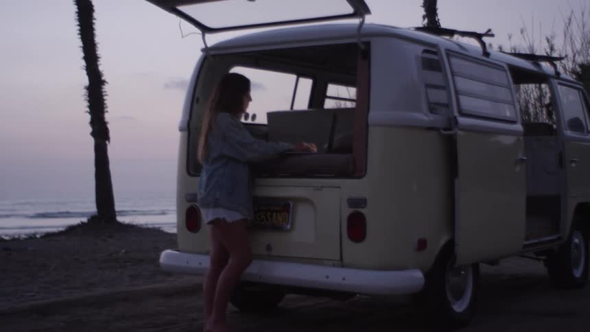 A woman stands at the back of her open van that is parked on the shore