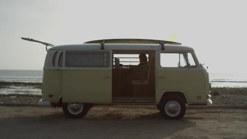 A woman sits in the back of her yellow van and types on a laptop while at the beach