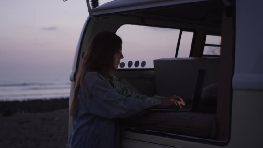A young woman stands behind her van that is parked on the beach and types on her laptop