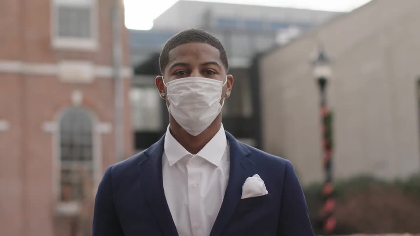 A young black man in a medical mask and a suit looks into the camera while standing outside of a large building during the day