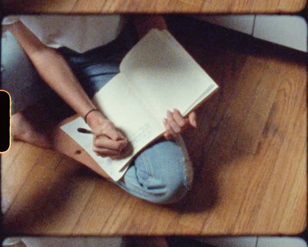 A person in a room sitting on the floor and writing in a book with a pen
