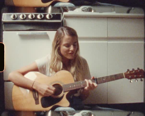 A woman sits on the floor and strums on her acoustic guitar while singing