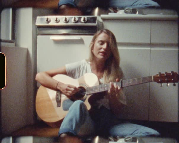 A blonde woman singing while playing an acoustic guitar on a kitchen floor