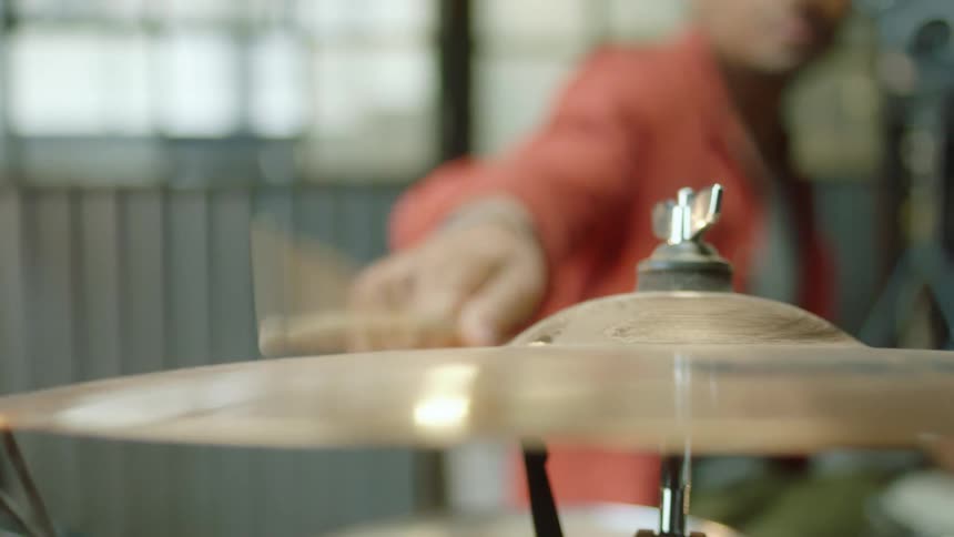 A person sitting in front of a drum set repeatedly strikes a cymbal with a drum stick before stopping the cymbals vibration with their hand