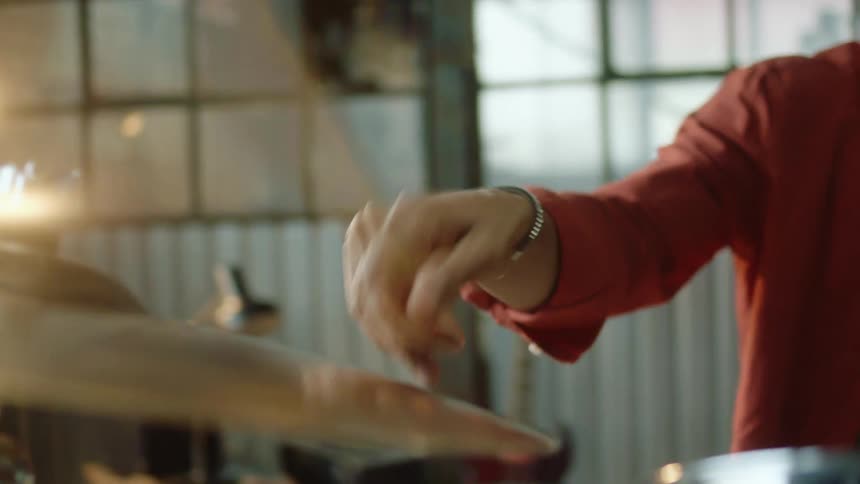 A drummer sitting before a drum set repeatedly strikes a cymbal with a drumstick indoors during the day