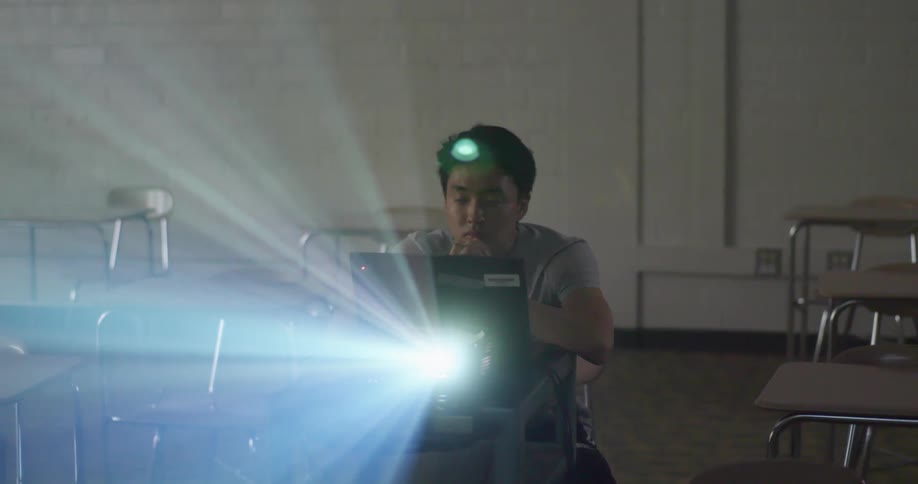 A teenage boy sits alone at a school desk with a laptop using a projector