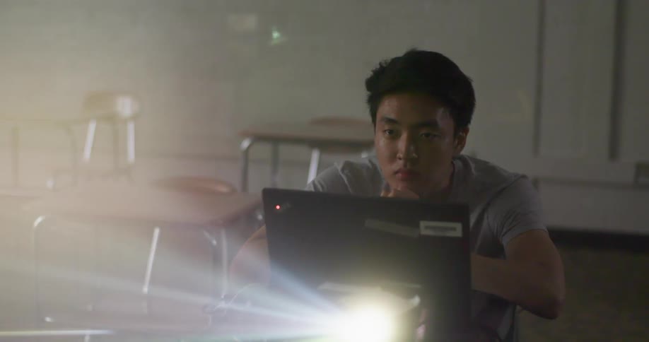 A teenage boy is sitting in a classroom by a laptop