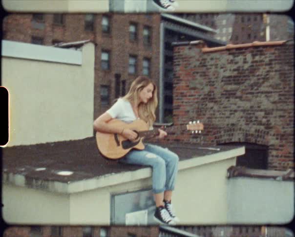 A woman sitting on a rooftop playing a guitar with buildings in the background