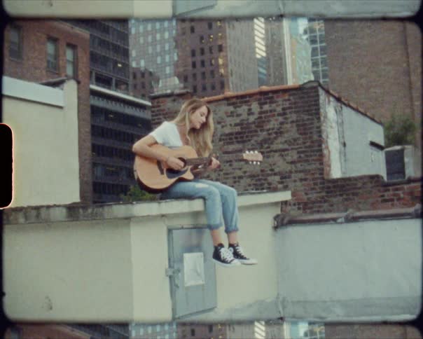 A young woman sings and plays the guitar while sitting on the rooftop of a building in a metropolis during the day