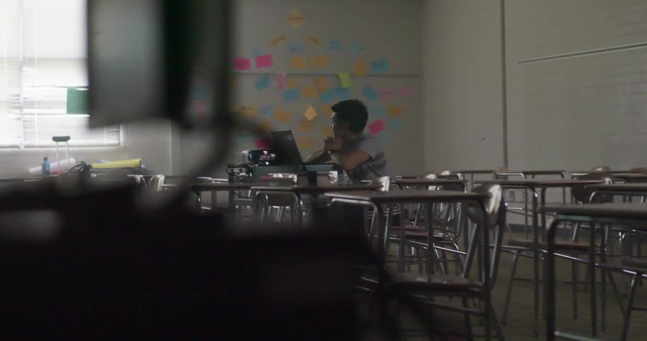 A lone boy sits in front of laptop at a desk in a school classroom during the day