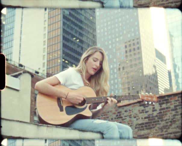 A woman is sitting in a large city and playing the guitar