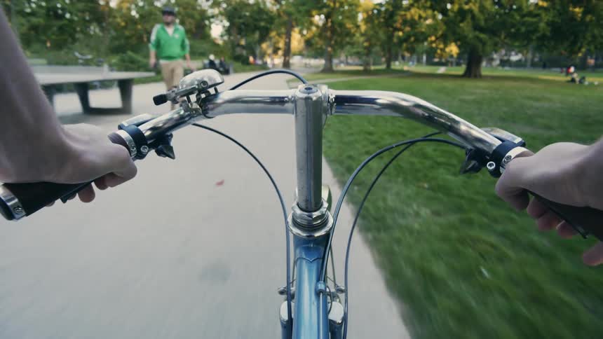 A man cycles through a public park filled with people on benches the path and the grass POV