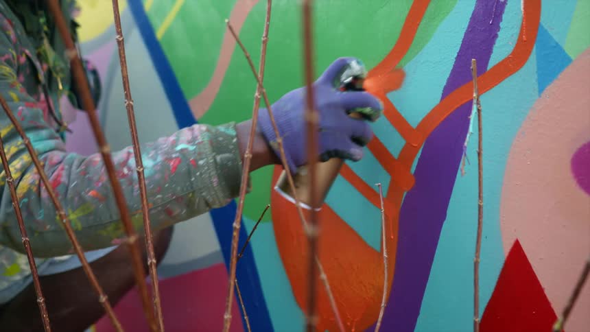 A man uses spray paint over a taped design on an outdoor mural