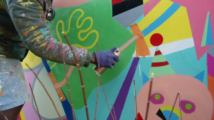 A painter uses a bottle of spray paint to paint a mural on a wall outdoors during the day