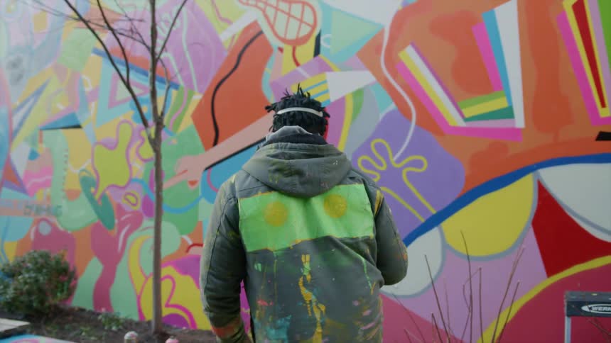 A man with a mask and gloves and a spray can walking up to a wall with graffiti