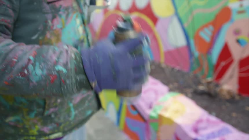 A person in paint covered clothing shakes a can of spray paint while standing outdoors during the day
