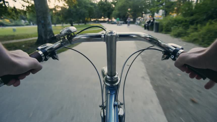 POV: A person rides a bike down a path in a public park with others riding skateboarding and sitting on a bench