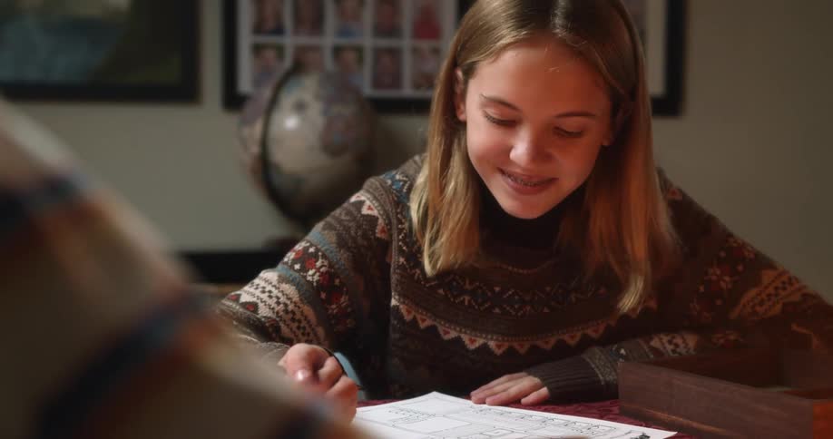 A girl with braces sitting at a desk writing on a paper with a globe and a wall in the background